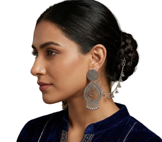 A woman in profile wearing large silver-toned traditional Indian jhumka earrings with dangling chain details, paired with a dark blue velvet top against a warm brown textured background.