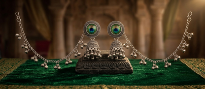 A pair of ornate silver-toned Indian jhumka earrings with peacock feather-inspired green and blue inlays, displayed on a carved black stand over a green velvet cloth with gold embroidery.