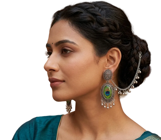 A woman in profile wearing large silver-toned statement earrings with peacock-eye motifs and chain details, paired with a teal sari and a braided updo.