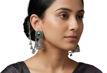 A close-up portrait of a woman wearing ornate silver-toned Indian jhumka earrings with circular green and blue stone centers and dangling bell-shaped charms, against a soft gray studio background.