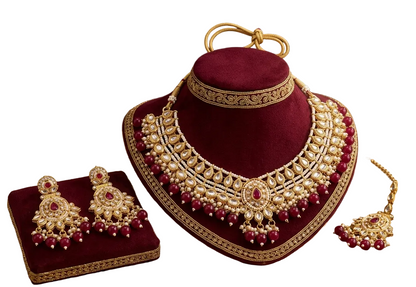 An ornate gold and deep red jewelry set displayed on velvet stands, with matching earrings on a small stand beside a necklace. The set includes a necklace, matching earrings, and a bracelet, styled on a patterned surface with decorative props and the text 'The Heritage Collection' in the upper left.