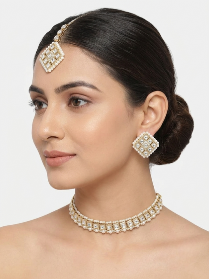 A woman with dark hair styled in a bun wears a matching gold-toned jewelry set featuring a choker necklace, a square-shaped headpiece, and matching earrings with clear stones.