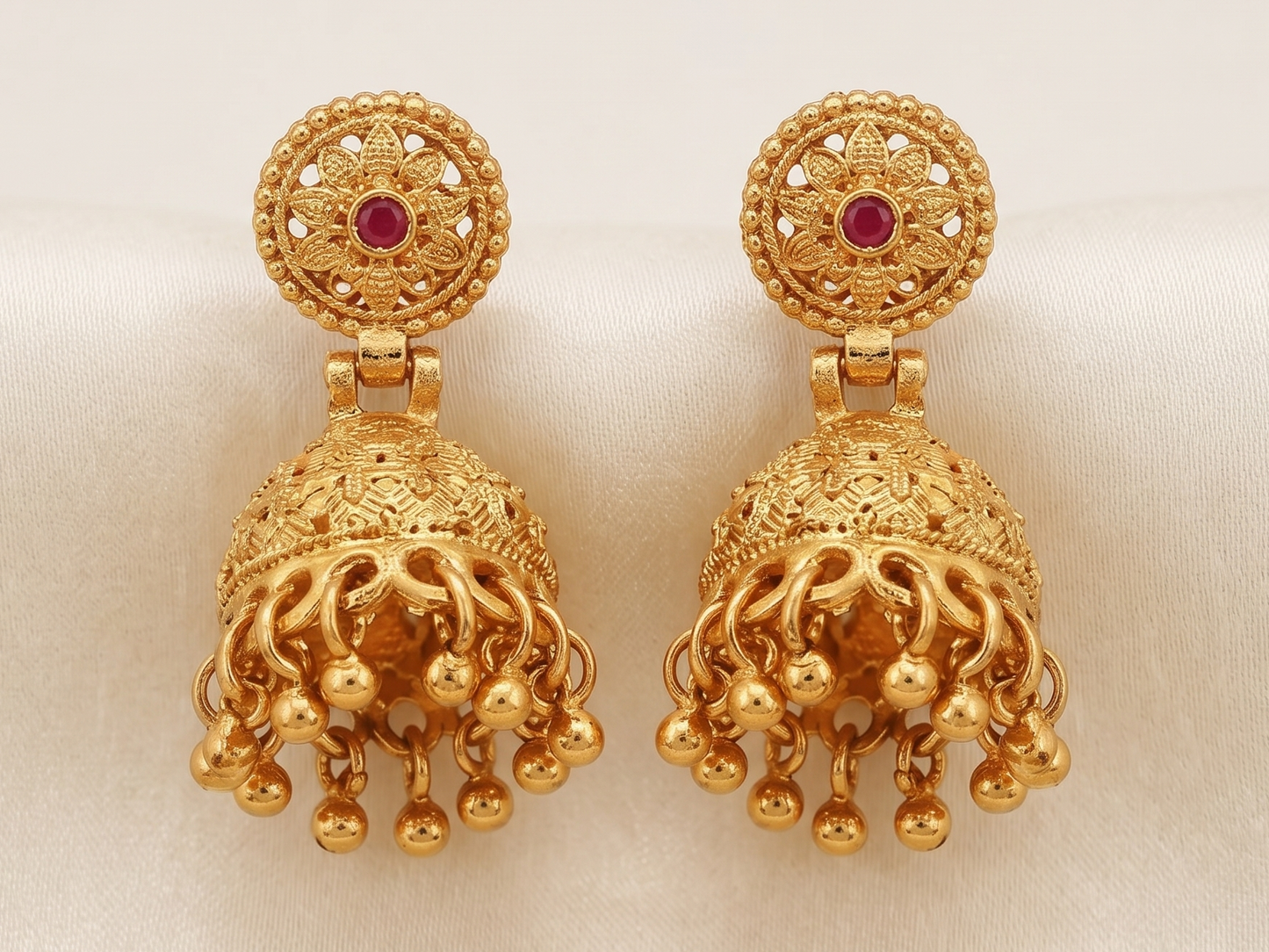 Close-up of Vaidika Royal Heritage gold-plated traditional Jhumka earrings featuring intricate temple design, a central red stone, and hanging golden bead drops resting on white silk fabric.