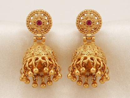 Close-up of Vaidika Royal Heritage gold-plated traditional Jhumka earrings featuring intricate temple design, a central red stone, and hanging golden bead drops resting on white silk fabric.