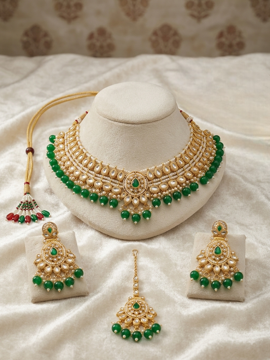 Gold and green traditional Indian jewelry set displayed on a cream-colored mannequin bust and small stands, including a necklace, earrings, and a dangling maang tikka, arranged on a white satin fabric background.