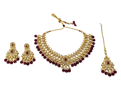 Gold-plated traditional Indian jewelry set featuring a multi-strand necklace with red bead accents, matching drop earrings, and a maang tikka, displayed on a solid white background.