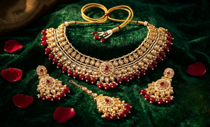 An ornate gold-toned traditional necklace set with matching earrings and a pendant, decorated with red beads and white stones, arranged on a dark green velvet fabric with scattered red rose petals.