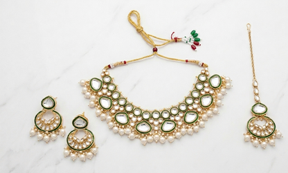 Gold-toned traditional jewelry set with a necklace, matching earrings, and a maang tikka, decorated with white faux pearls, green enamel, and clear stones, displayed on a white marble surface.
