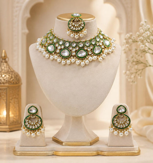 An ornate Indian-style jewelry set displayed on cream-colored mannequin stands, featuring a choker necklace, matching earrings, and a small matching piece on a stand. The jewelry is decorated with green stones, white pearls, and gold-toned metal, set against a warm, softly blurred background with traditional decor.
