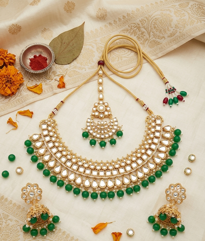 Gold-toned traditional Indian jewelry set with green bead accents, including a necklace, matching earrings, and a maang tikka, arranged on a cream patterned fabric with decorative flowers and a small bowl of red powder.