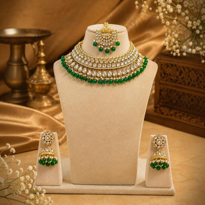 Gold and green traditional Indian jewelry set displayed on a beige velvet mannequin bust, including a multi-layered choker necklace, matching earrings, and a hair ornament, set against a warm golden background with decorative props.