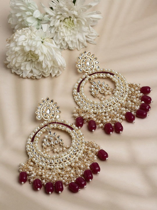 A pair of ornate Indian jhumka earrings with gold-toned metalwork, white faux pearls, and deep red bead accents, displayed on a beige fabric background with white flowers.