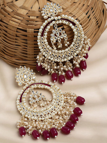 A pair of ornate Indian jhumka earrings with cream-colored faux pearls, silver-toned metalwork, and deep red bead drops, displayed on a beige fabric surface next to a woven basket.
